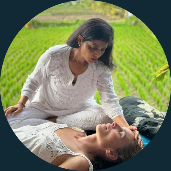 Woman is facilitating a breathwork retreat, you see a woman on a yoga mat and the facilitator softly placing a hand on her forehead